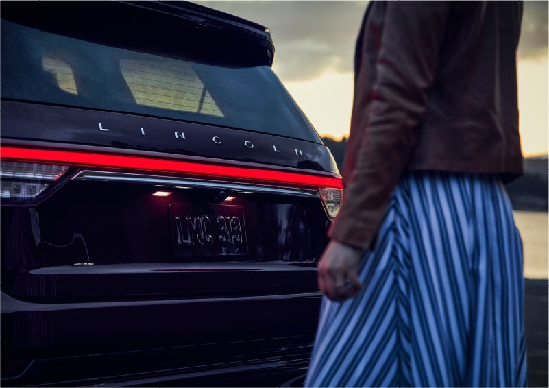 A person is shown near the rear of a 2023 Lincoln Aviator® SUV as the Lincoln Embrace illuminates the rear lights | Jenkins & Wynne Lincoln in Clarksville TN